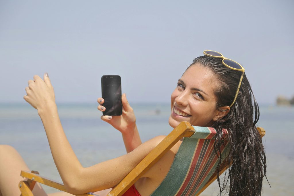 Smiling woman enjoying her vacation at Mgarr, Malta beach with a smartphone in hand.